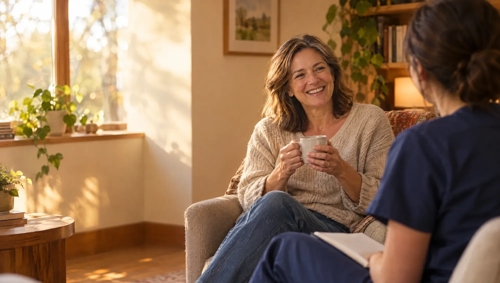 A woman in her fifties laughs over a cup of tea as a clinician listens, in a sunlit room