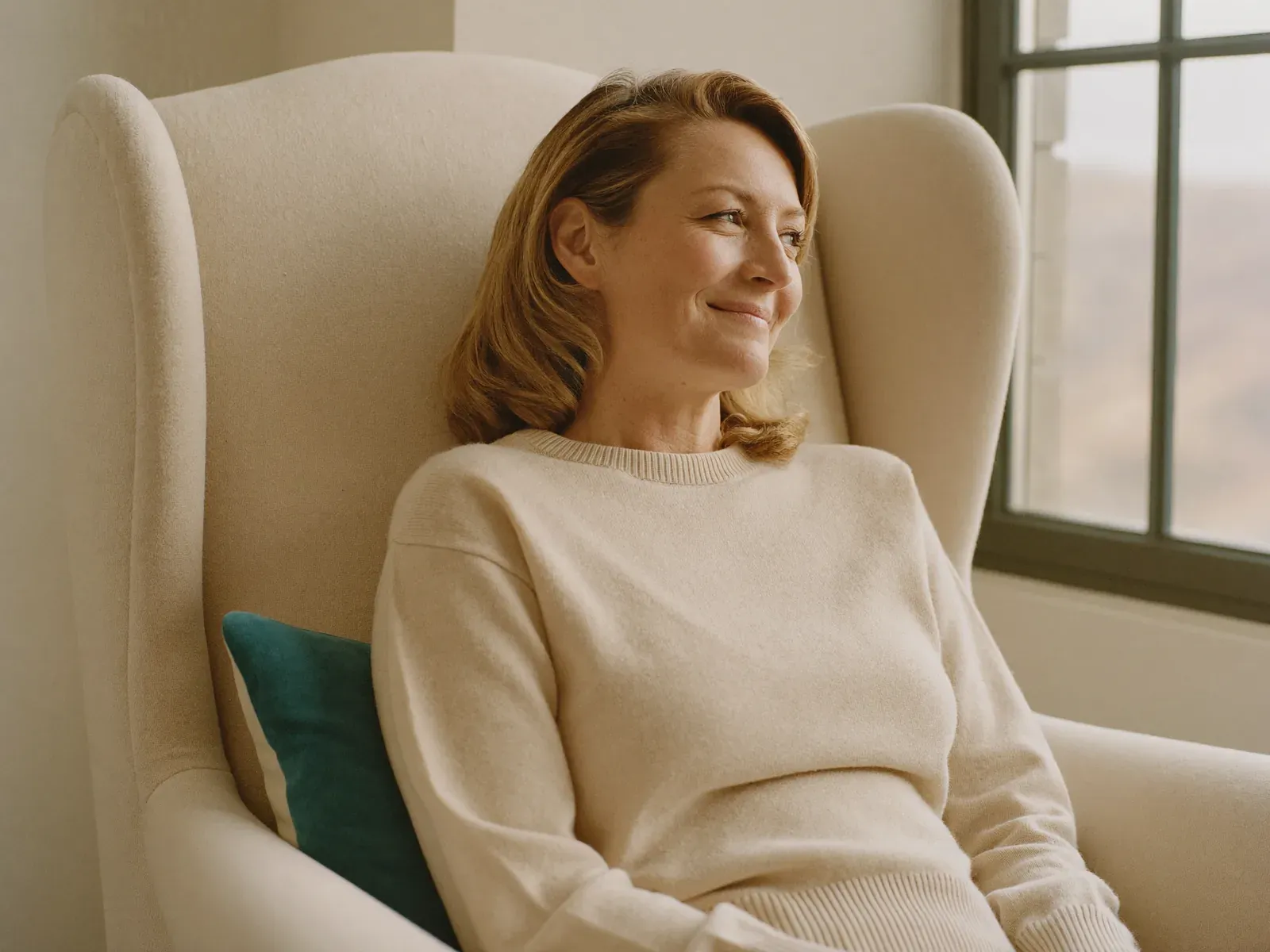 A woman in a cream sweater resting in an armchair by the window, smiling